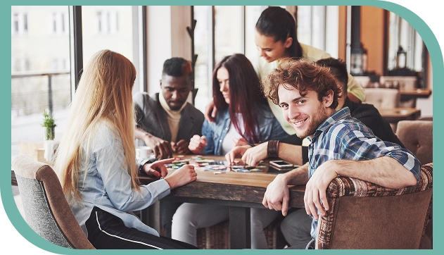 Een groep vrienden speelt gezellig een bordspel; een van de spelers kijkt naar de camera.